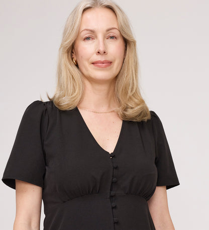 A woman with shoulder-length blonde hair wears the Maryam Black Button Up V-Neck Short Sleeve Top, standing against a plain light background and looking at the camera with a neutral expression.