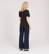 A woman with blonde hair, wearing the Maryam Black Button Up V-Neck Short Sleeve Top, dark wide-leg jeans, and black heels, stands facing away from the camera against a plain white background.
