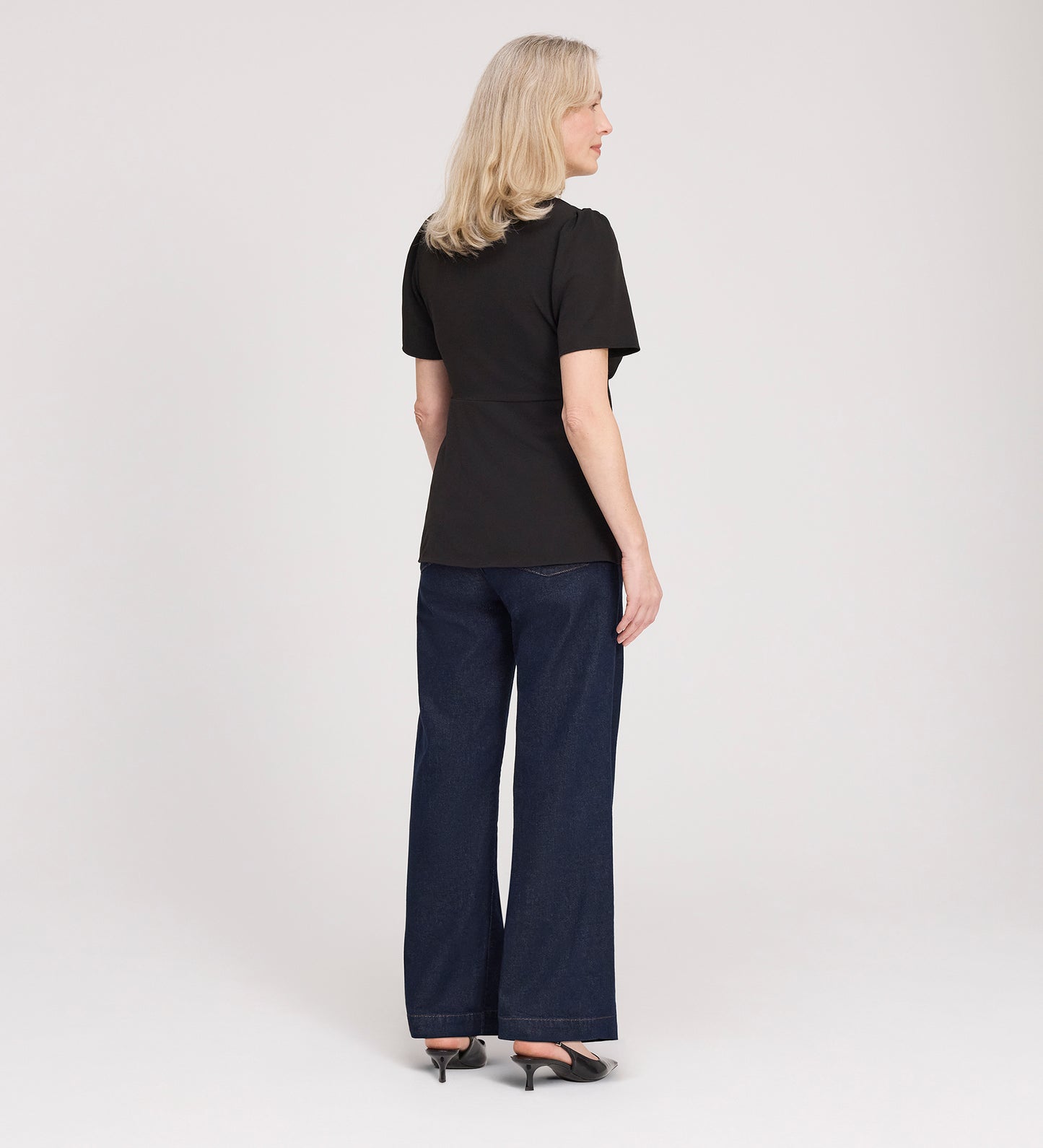 A woman with blonde hair, wearing the Maryam Black Button Up V-Neck Short Sleeve Top, dark wide-leg jeans, and black heels, stands facing away from the camera against a plain white background.