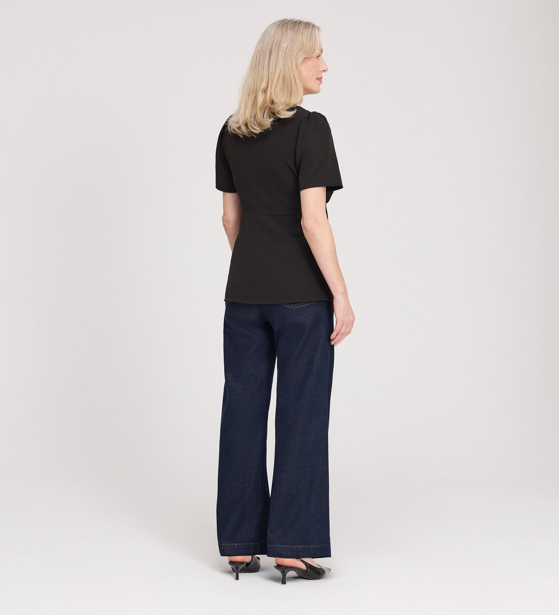 A woman with blonde hair, wearing the Maryam Black Button Up V-Neck Short Sleeve Top, dark wide-leg jeans, and black heels, stands facing away from the camera against a plain white background.