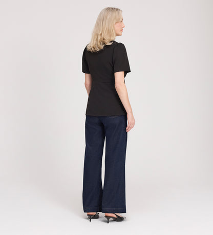 A woman with blonde hair, wearing the Maryam Black Button Up V-Neck Short Sleeve Top, dark wide-leg jeans, and black heels, stands facing away from the camera against a plain white background.