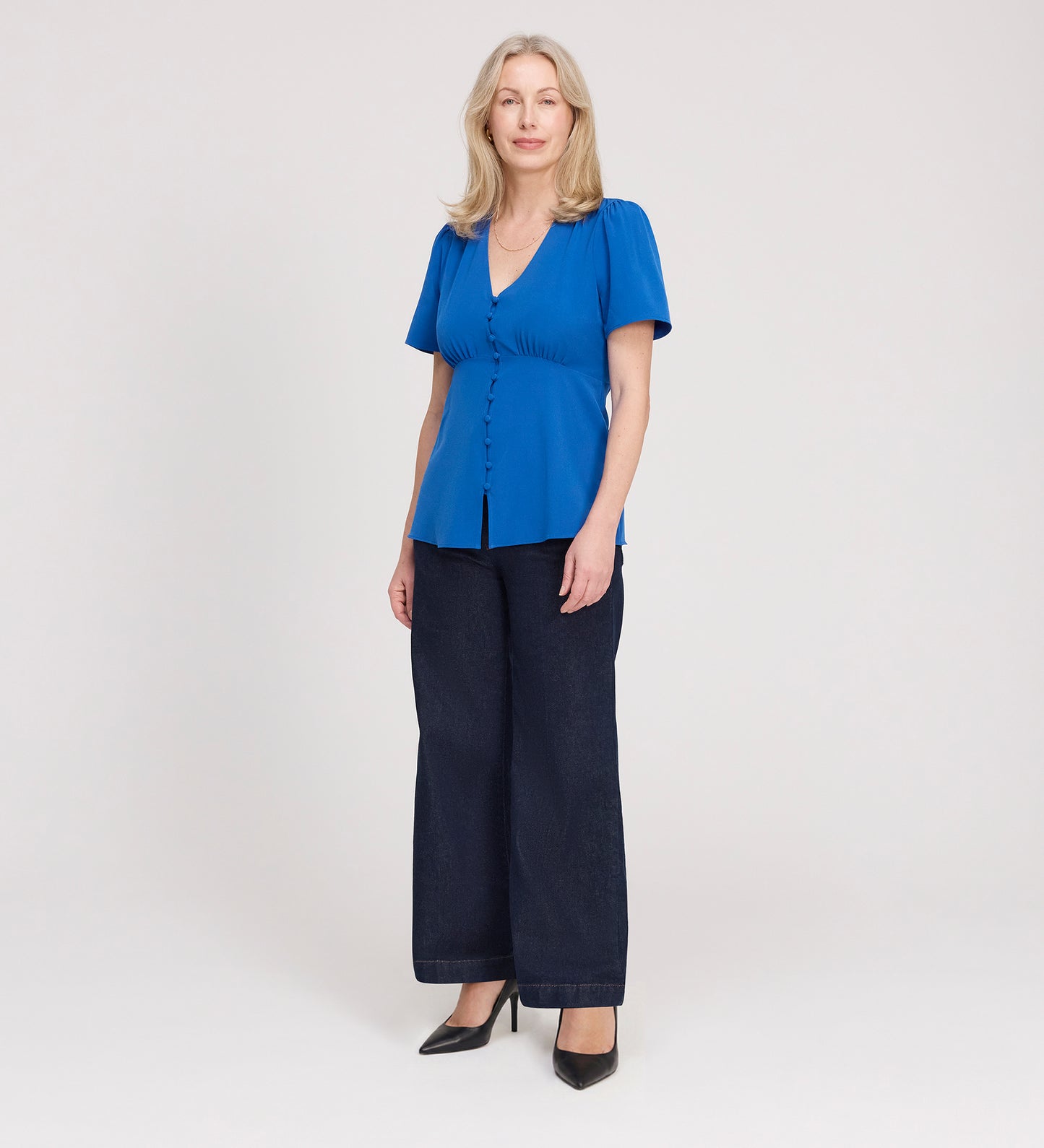 A woman with light hair stands against a plain background, facing forward with a neutral expression. She wears the Maryam Cobalt Button Up V-Neck Short Sleeve Top, dark wide-leg jeans, and black heels.