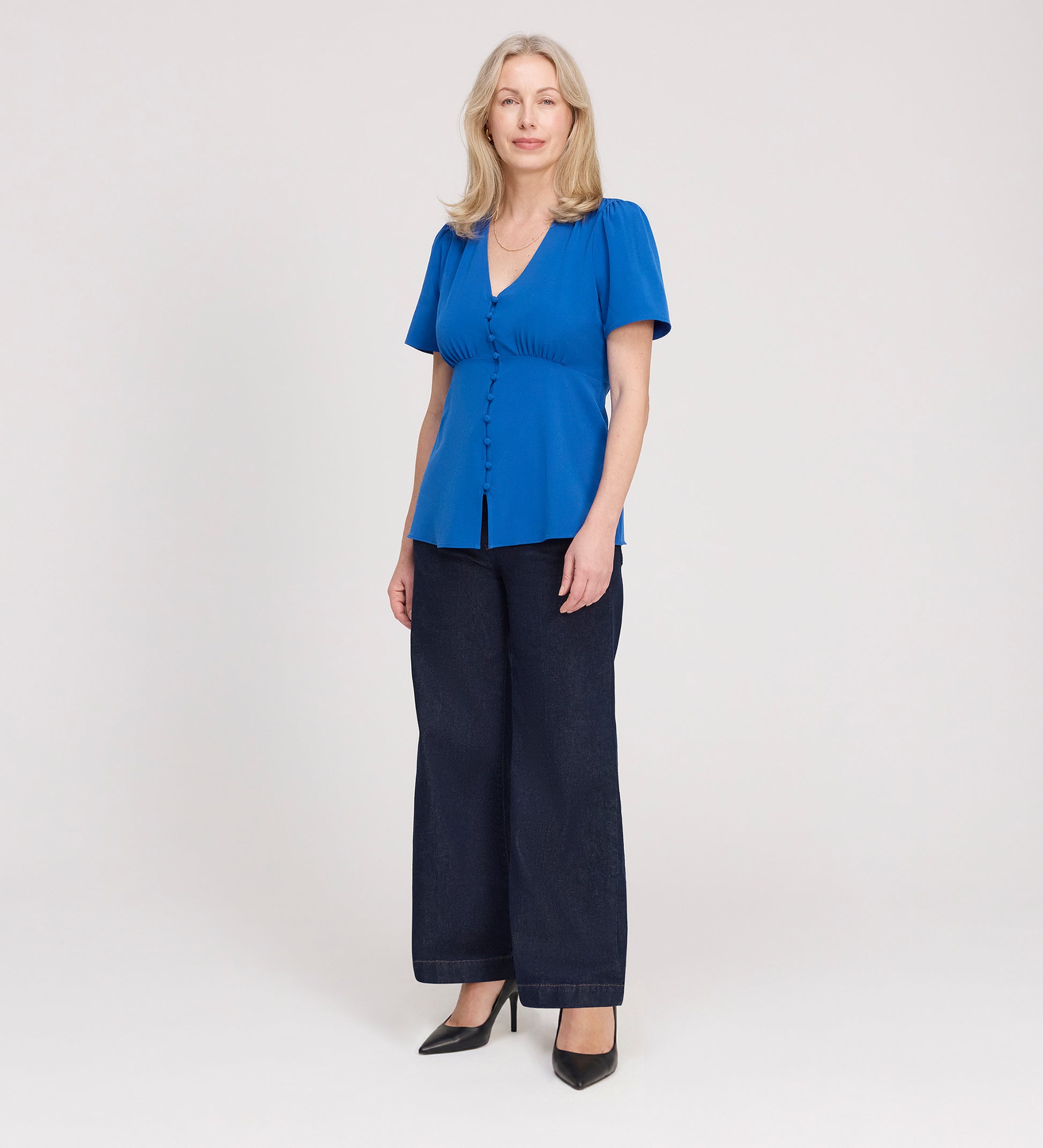 A woman with light hair stands against a plain background, facing forward with a neutral expression. She wears the Maryam Cobalt Button Up V-Neck Short Sleeve Top, dark wide-leg jeans, and black heels.