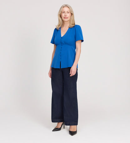 A woman with light hair stands against a plain background, facing forward with a neutral expression. She wears the Maryam Cobalt Button Up V-Neck Short Sleeve Top, dark wide-leg jeans, and black heels.