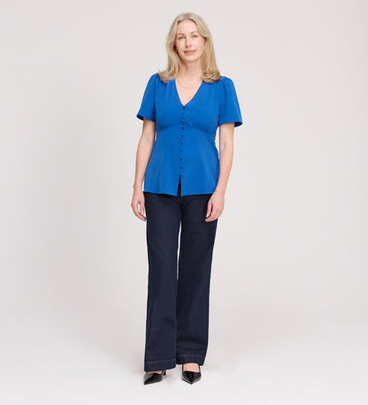 A woman with long blonde hair faces forward, wearing the Maryam Cobalt Button Up V-Neck Short Sleeve Top, dark wide-leg pants, and black pointed shoes against a light, plain background.