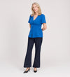A woman with light hair stands against a plain background, wearing the Maryam Cobalt Button Up V-Neck Short Sleeve Top, dark pants, and black heels. She smiles softly with her hands behind her back.