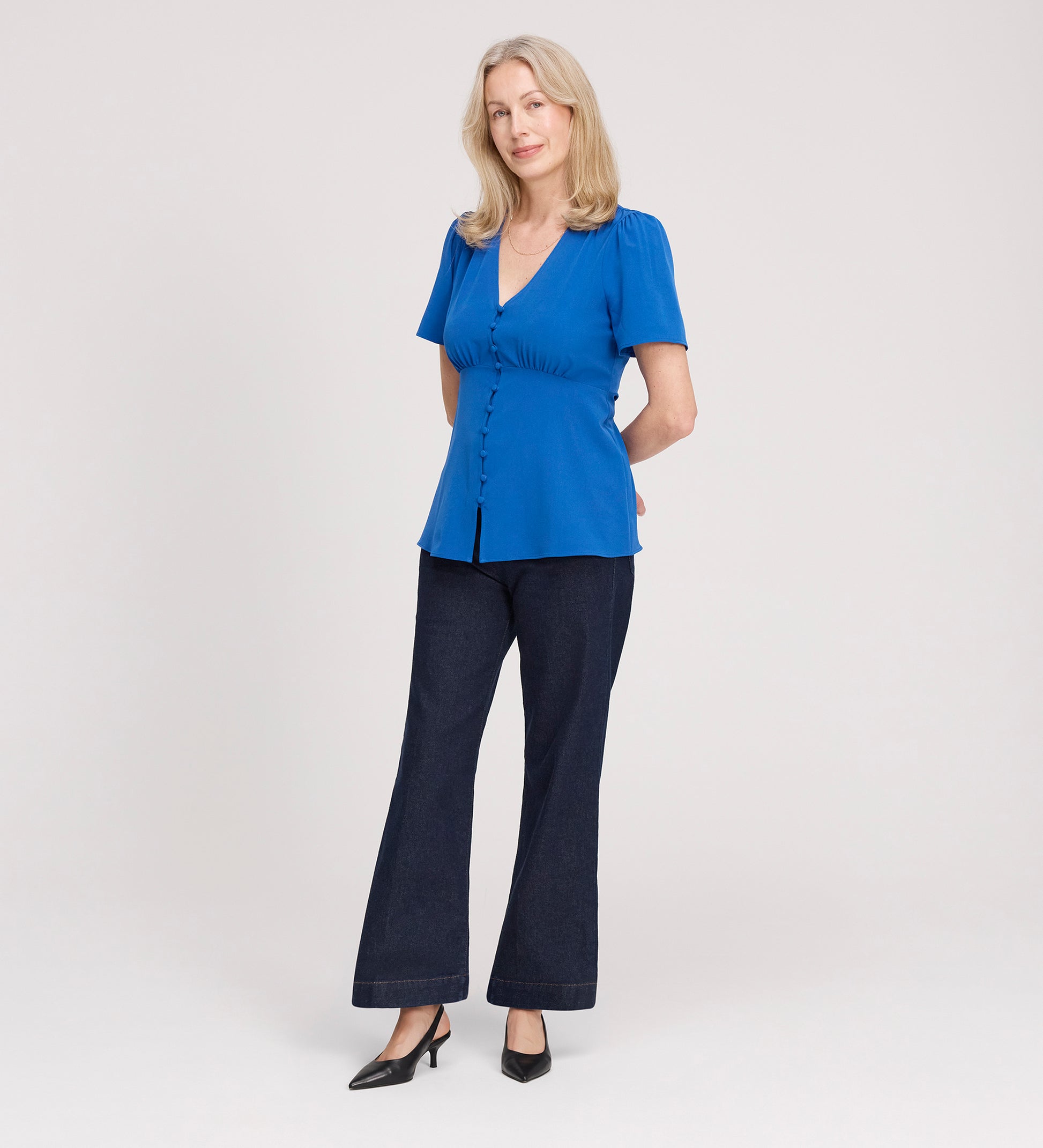 A woman with light hair stands against a plain background, wearing the Maryam Cobalt Button Up V-Neck Short Sleeve Top, dark pants, and black heels. She smiles softly with her hands behind her back.