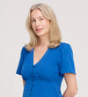 A woman with shoulder-length blonde hair smiles softly at the camera, wearing a Maryam Cobalt Button Up V-Neck Short Sleeve Top and standing against a plain light background.