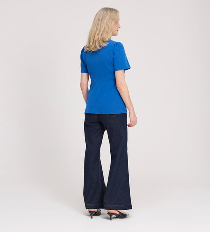 A woman with blonde hair faces away, wearing a Maryam Cobalt Button Up V-Neck Short Sleeve Top, dark flared jeans, and black heels, set against a plain white background.