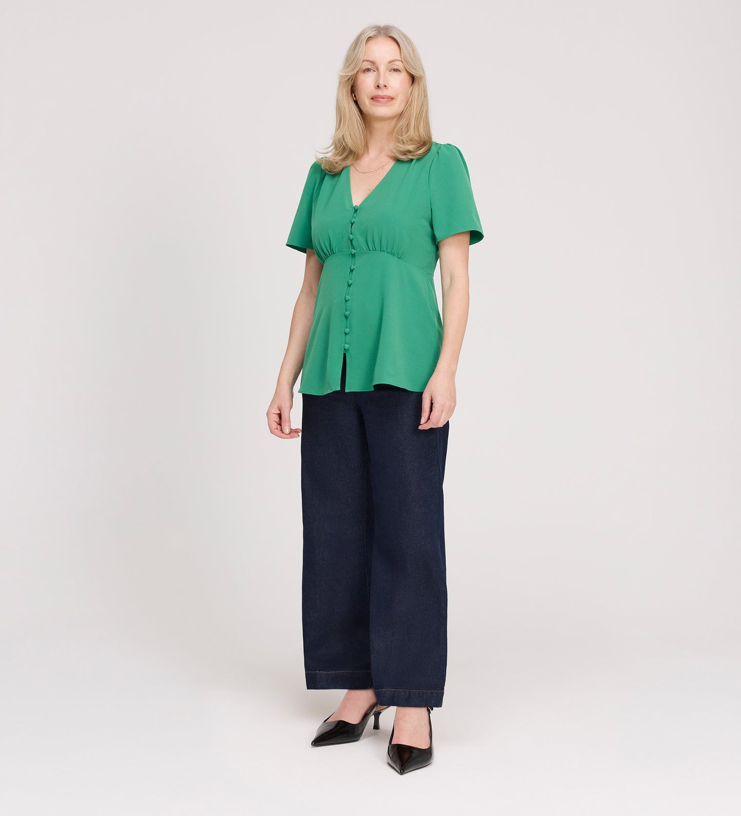 A woman with light hair stands against a white background, wearing the Maryam Green Button Up V-Neck Short Sleeve Top and dark wide-leg pants. She pairs the blouse with black pointed shoes and looks at the camera with a slight smile.