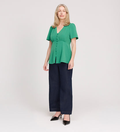A woman with light hair stands against a white background, wearing the Maryam Green Button Up V-Neck Short Sleeve Top and dark wide-leg pants. She pairs the blouse with black pointed shoes and looks at the camera with a slight smile.