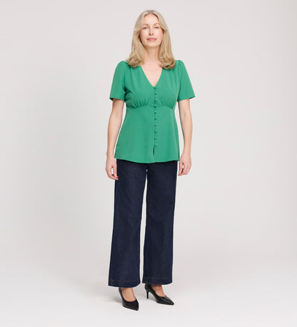 A woman with long blonde hair faces forward, wearing the Maryam Green Button Up V-Neck Short Sleeve Top, dark wide-leg pants, and black heels against a plain light background.