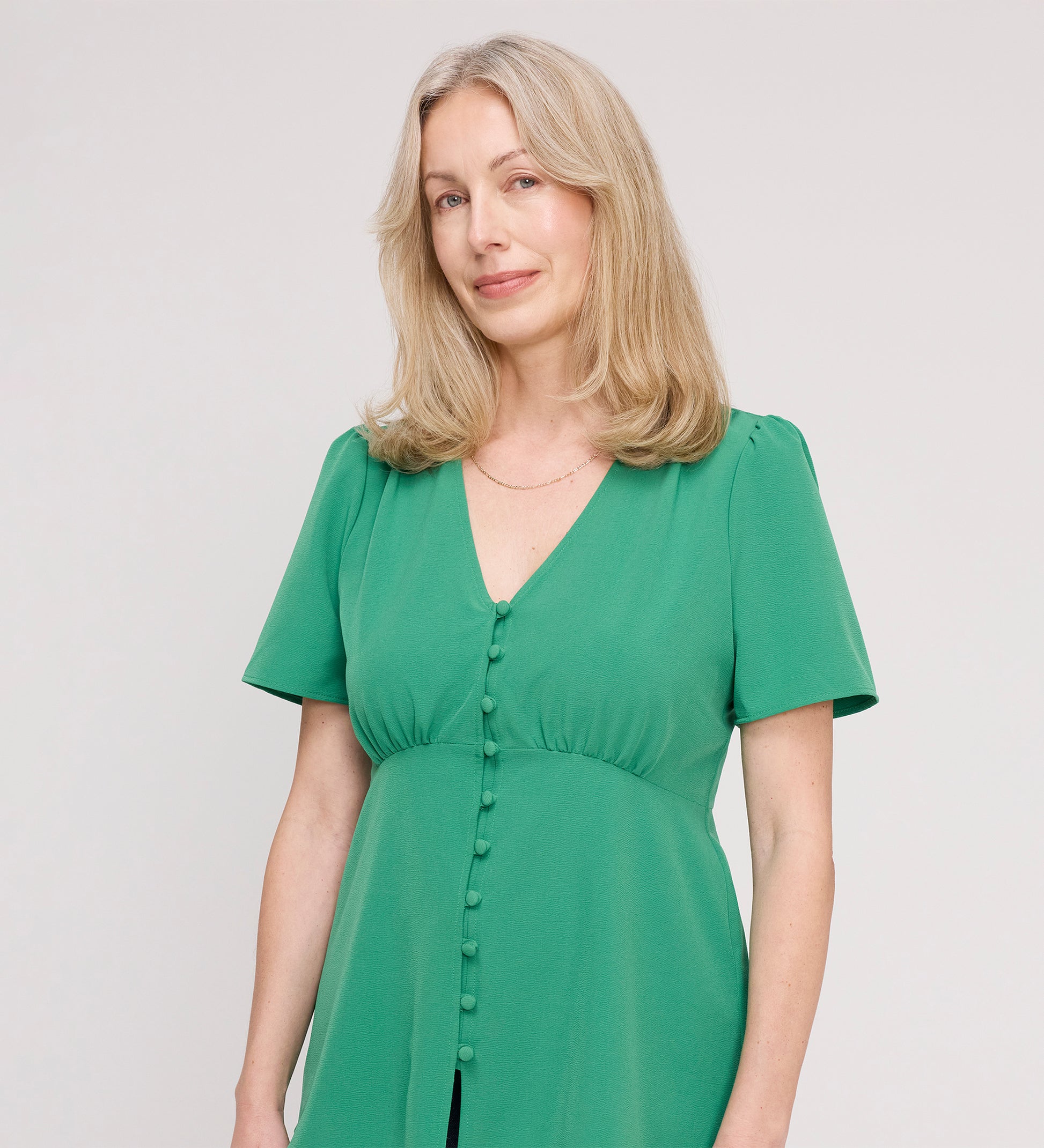 A woman with shoulder-length blonde hair wears the Maryam Green Button Up V-Neck Short Sleeve Top, smiling softly against a plain light background.