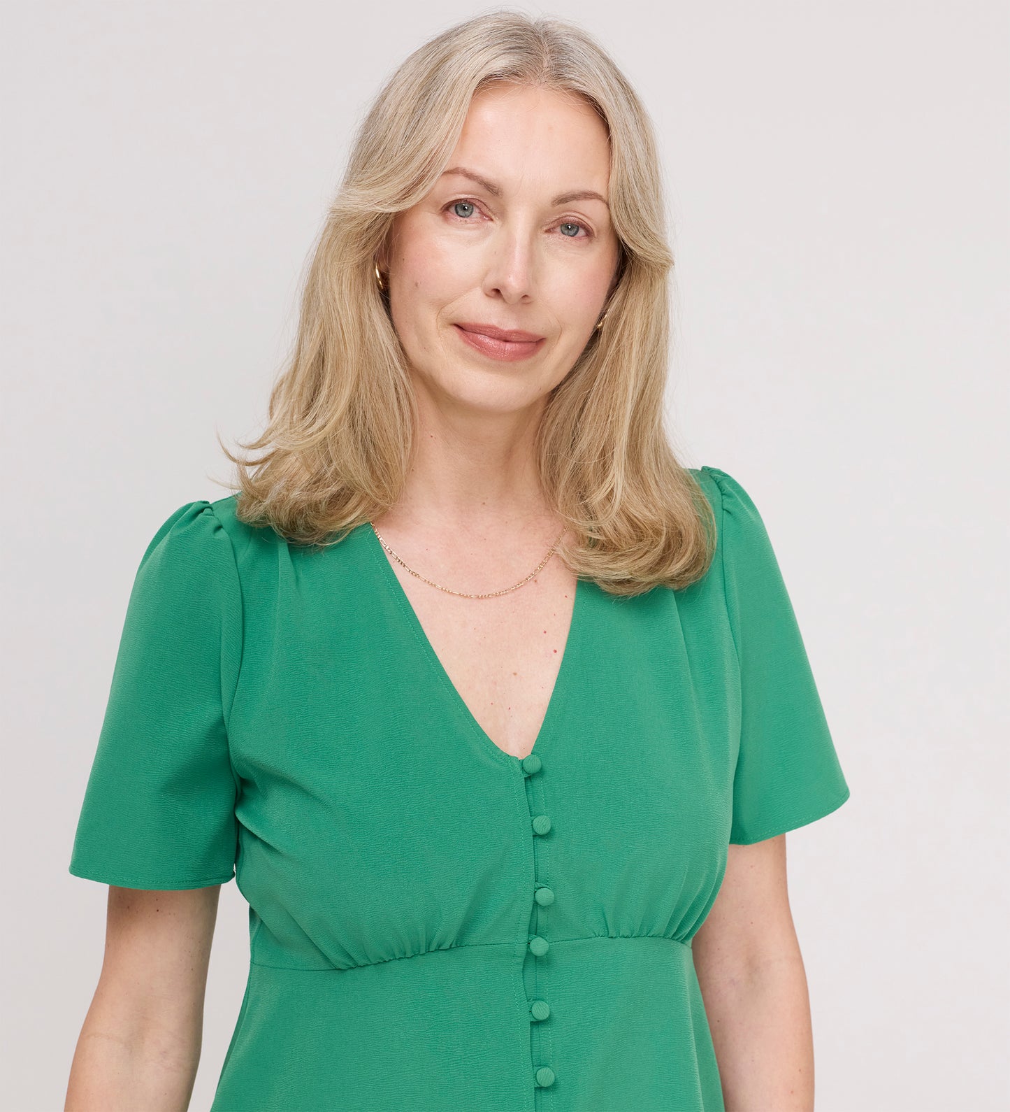 A woman with light blonde hair smiles gently at the camera, wearing the Maryam Green Button Up V-Neck Short Sleeve Top against a plain, light background.
