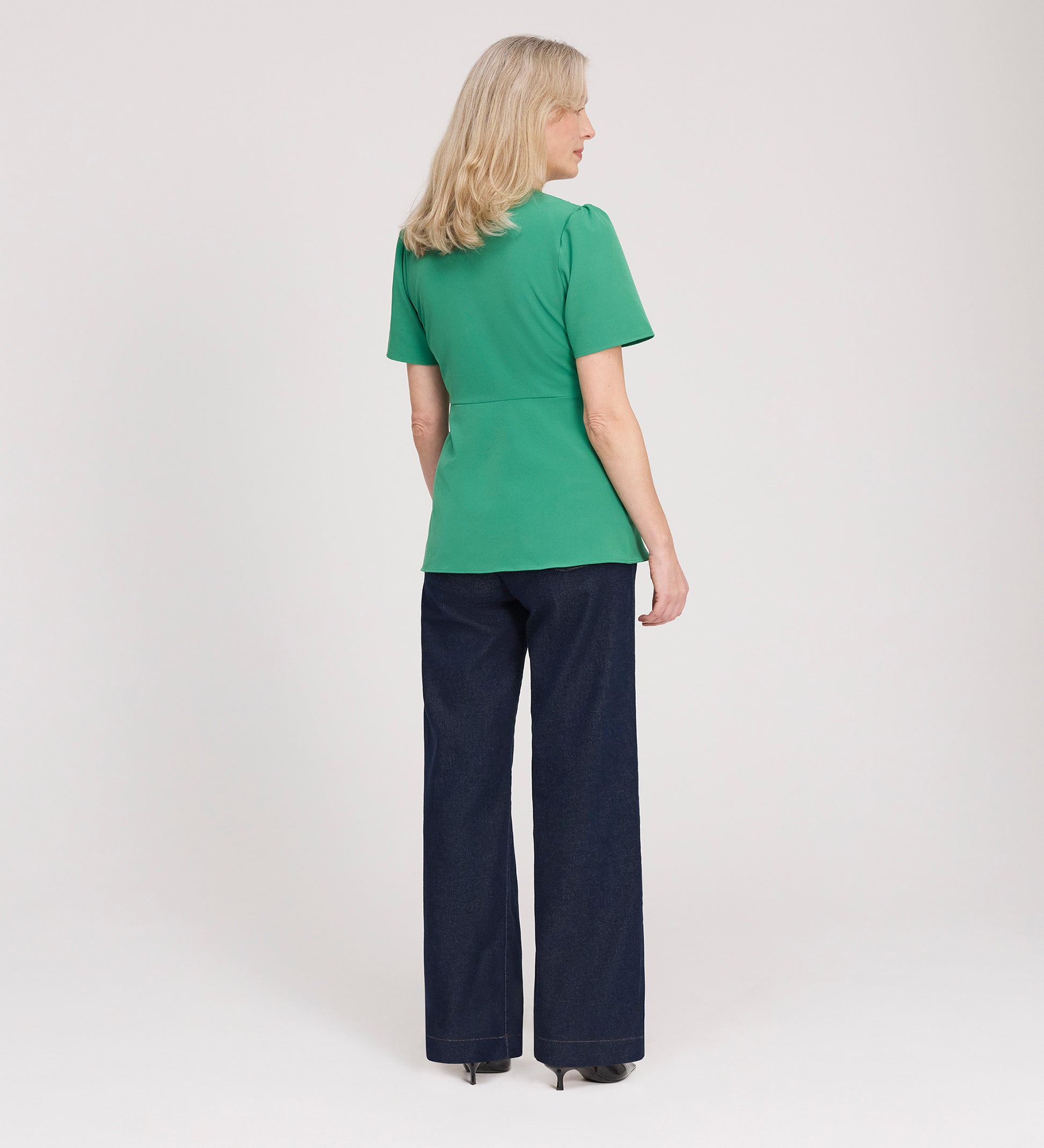 A woman with blonde hair faces away, wearing the Maryam Green Button Up V-Neck Short Sleeve Top, dark wide-leg pants, and heels against a plain white background.