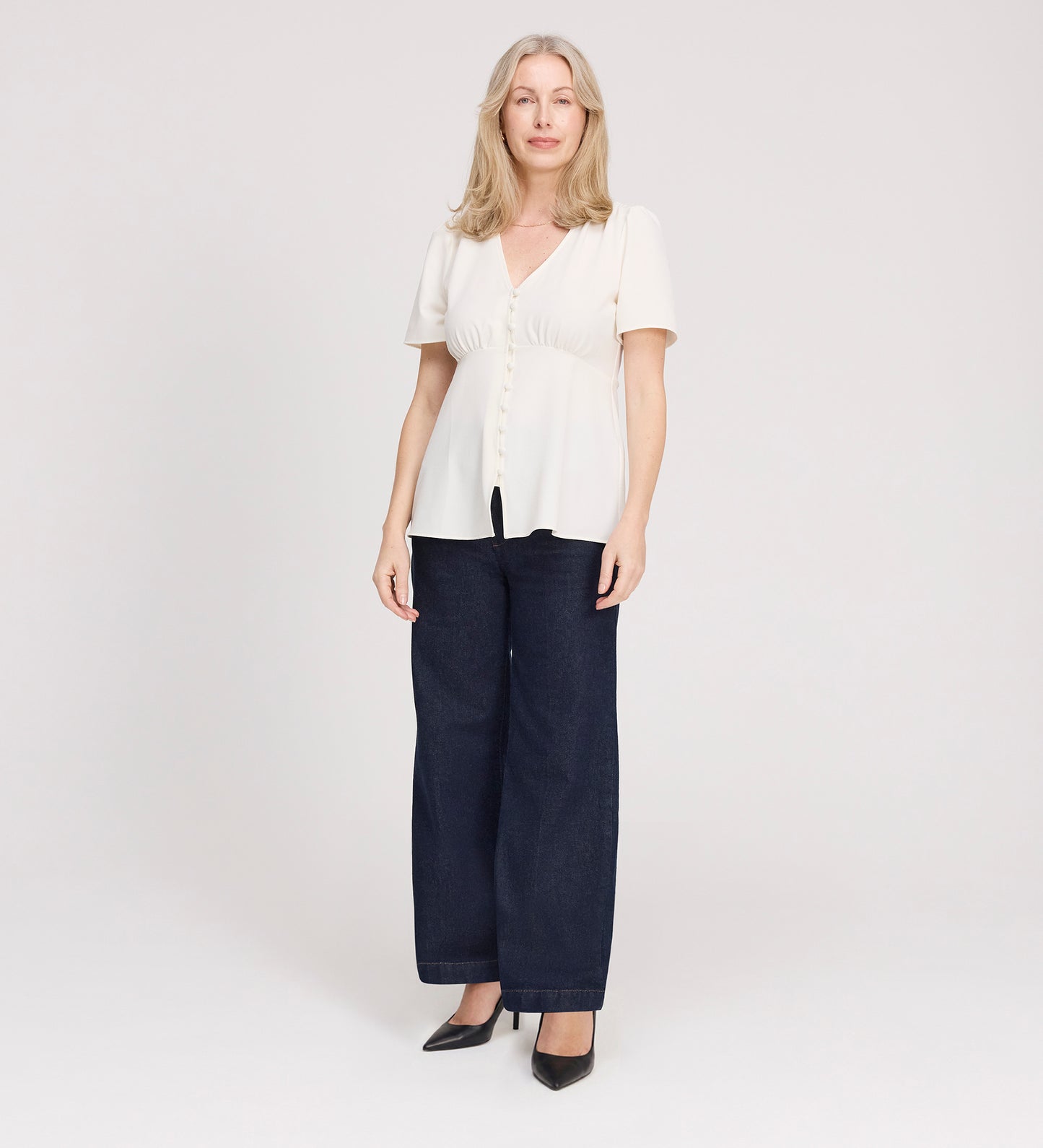 A woman with light blond hair wears the Maryam Ivory Button Up V-Neck Short Sleeve Top, paired with dark wide-leg jeans and black heels, standing facing forward against a plain white background.