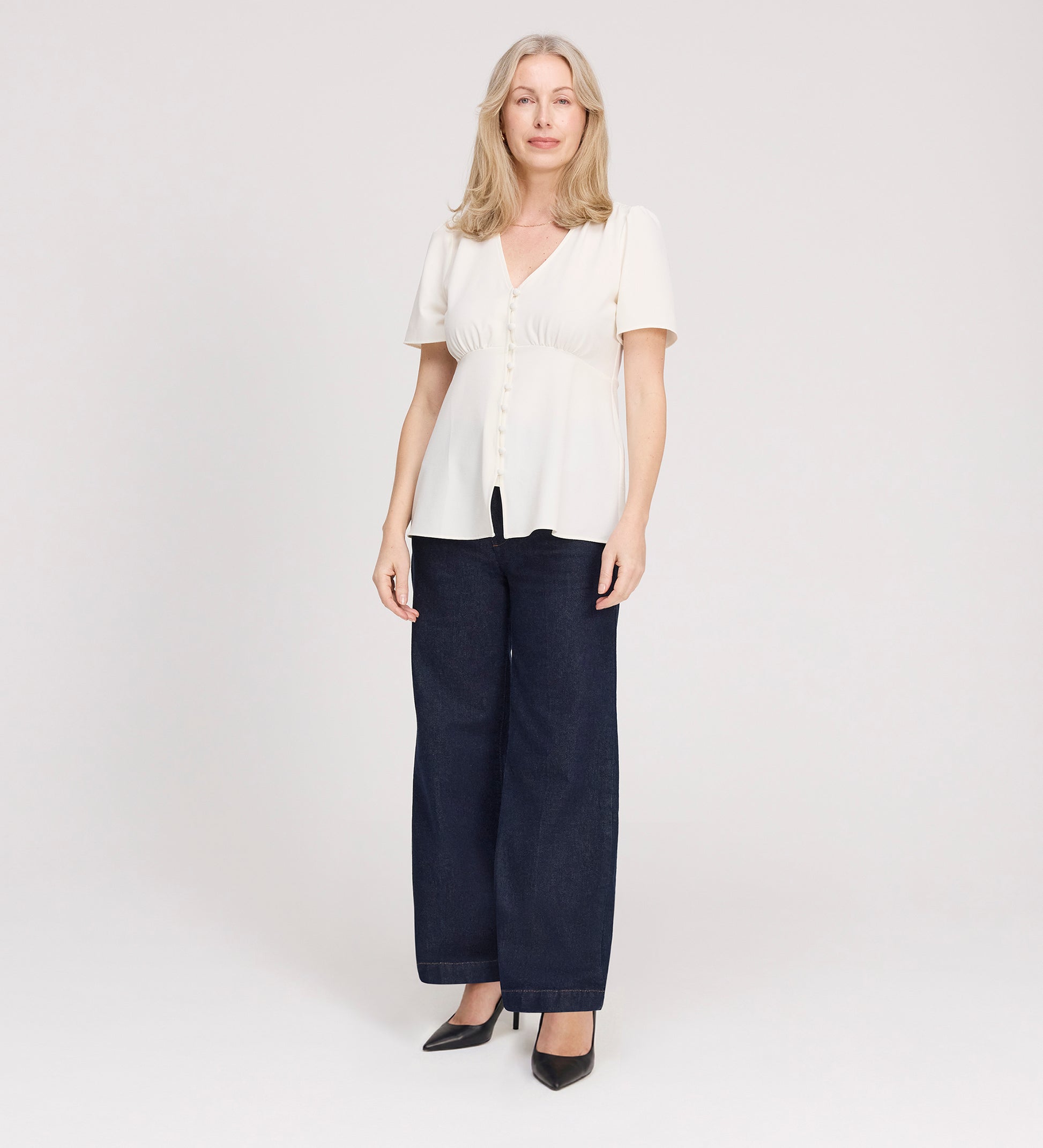 A woman with light blond hair wears the Maryam Ivory Button Up V-Neck Short Sleeve Top, paired with dark wide-leg jeans and black heels, standing facing forward against a plain white background.