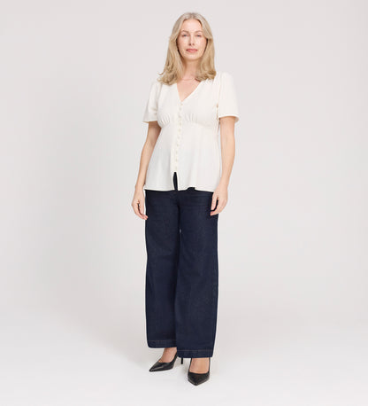 A woman with light blond hair wears the Maryam Ivory Button Up V-Neck Short Sleeve Top, paired with dark wide-leg jeans and black heels, standing facing forward against a plain white background.