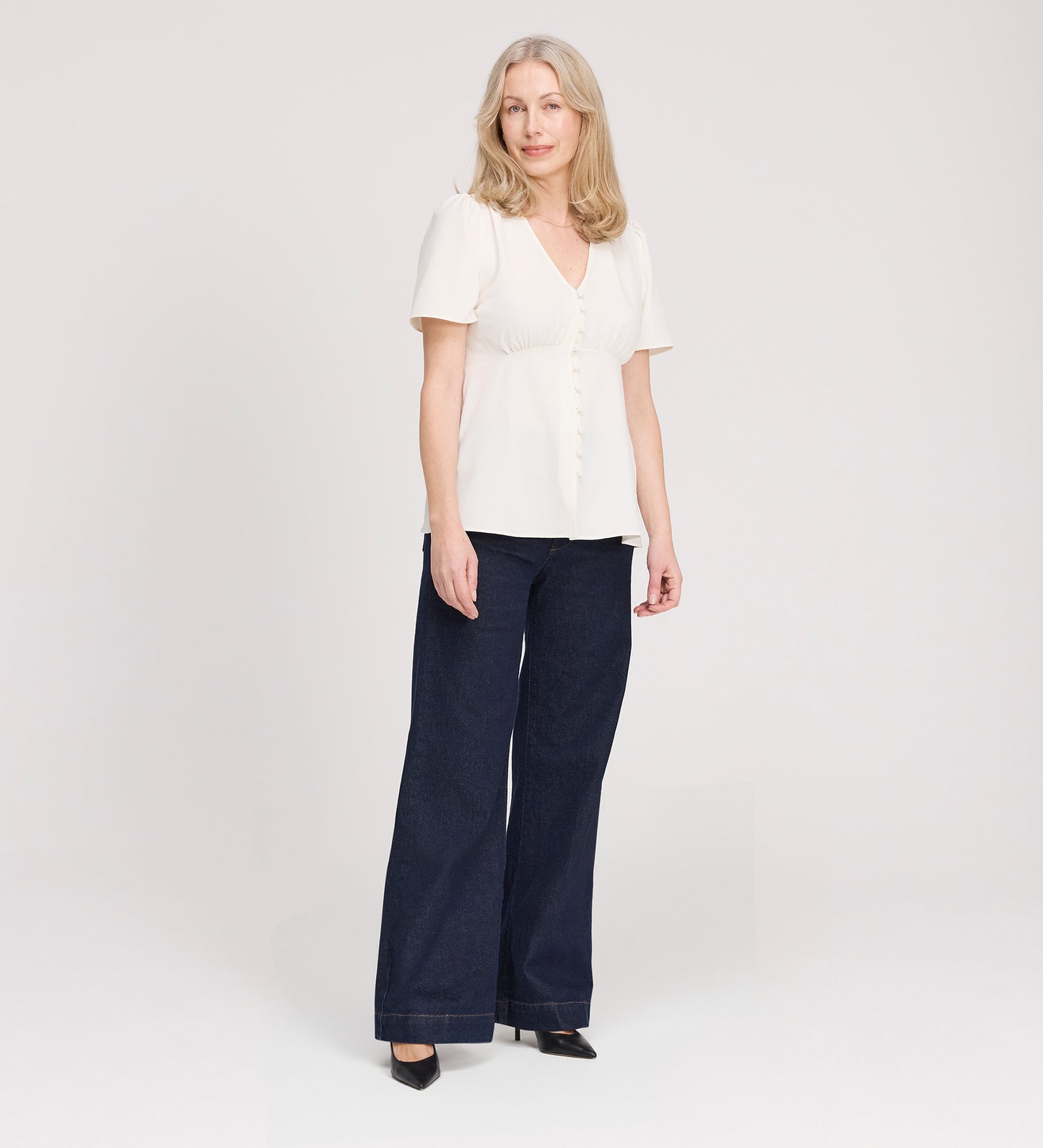 A woman with light hair wears the Maryam Ivory Button Up V-Neck Short Sleeve Top with dark wide-leg pants and black heels, standing against a plain light background. Her expression is relaxed and neutral.
