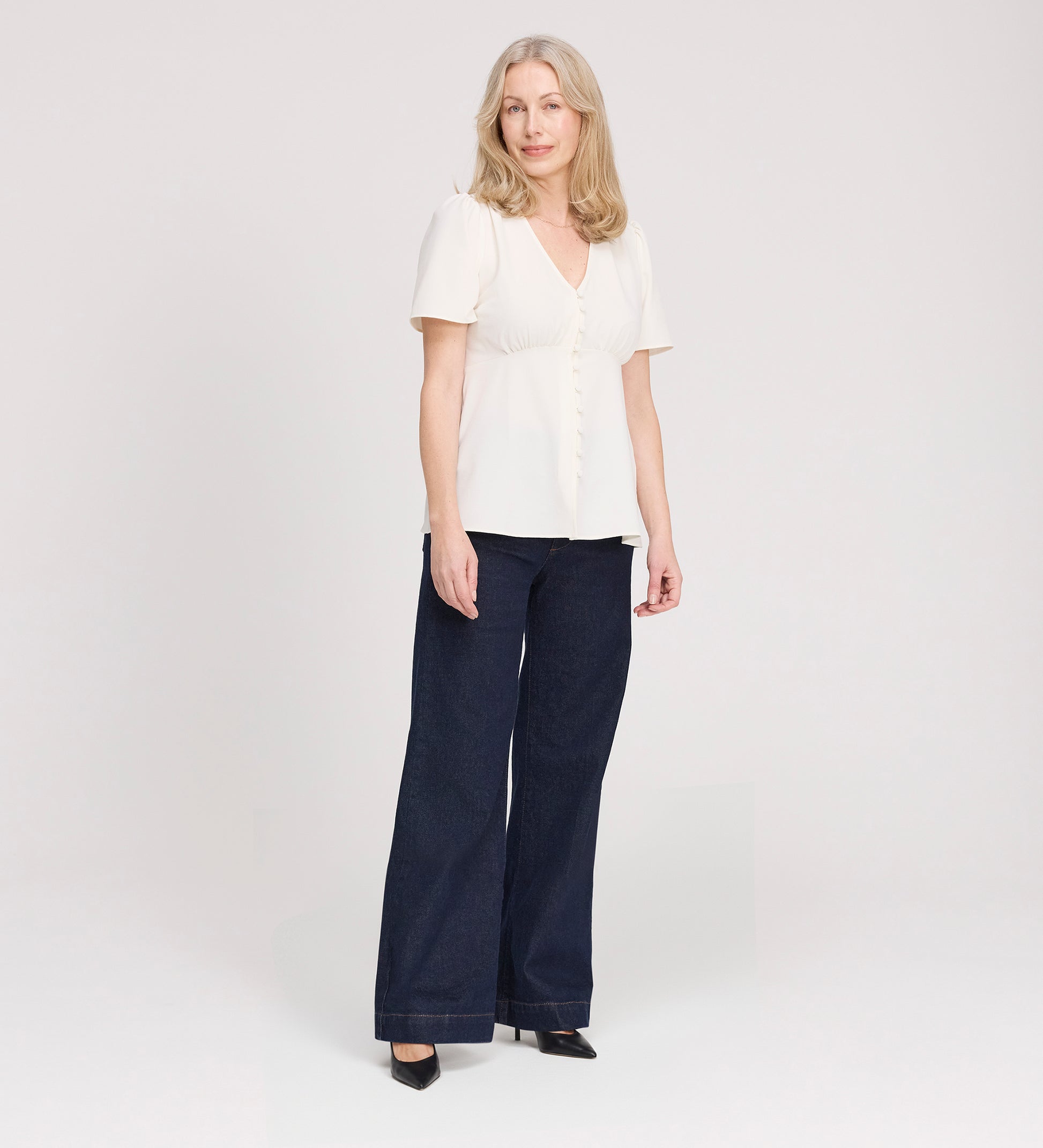 A woman with light hair wears the Maryam Ivory Button Up V-Neck Short Sleeve Top with dark wide-leg pants and black heels, standing against a plain light background. Her expression is relaxed and neutral.