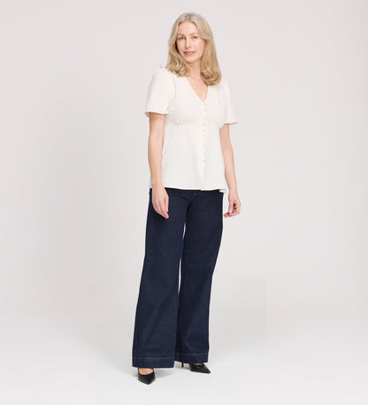 A woman with light hair wears the Maryam Ivory Button Up V-Neck Short Sleeve Top with dark wide-leg pants and black heels, standing against a plain light background. Her expression is relaxed and neutral.