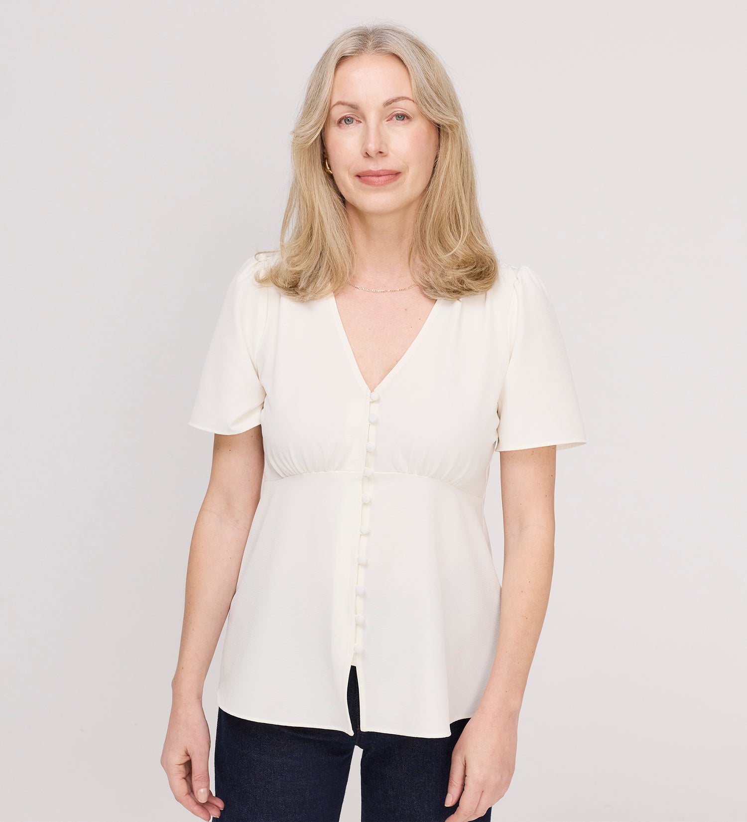 A woman with shoulder-length blonde hair smiles softly as she stands against a plain light background, wearing the Maryam Ivory Button Up V-Neck Short Sleeve Top paired with dark pants.