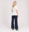 A woman with shoulder-length blonde hair faces away, wearing the Maryam Ivory Button Up V-Neck Short Sleeve Top, dark wide-leg jeans, and black heels against a plain light background.