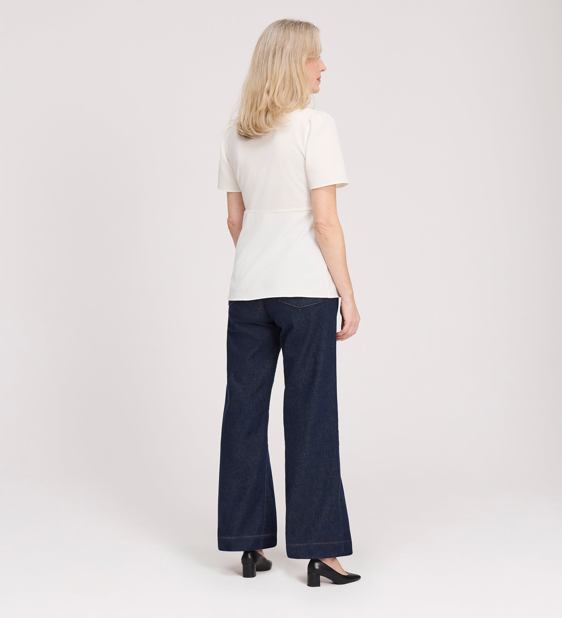 A woman with shoulder-length blonde hair faces away, wearing the Maryam Ivory Button Up V-Neck Short Sleeve Top, dark wide-leg jeans, and black heels against a plain light background.
