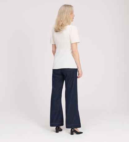 A woman with shoulder-length blonde hair faces away, wearing the Maryam Ivory Button Up V-Neck Short Sleeve Top, dark wide-leg jeans, and black heels against a plain light background.