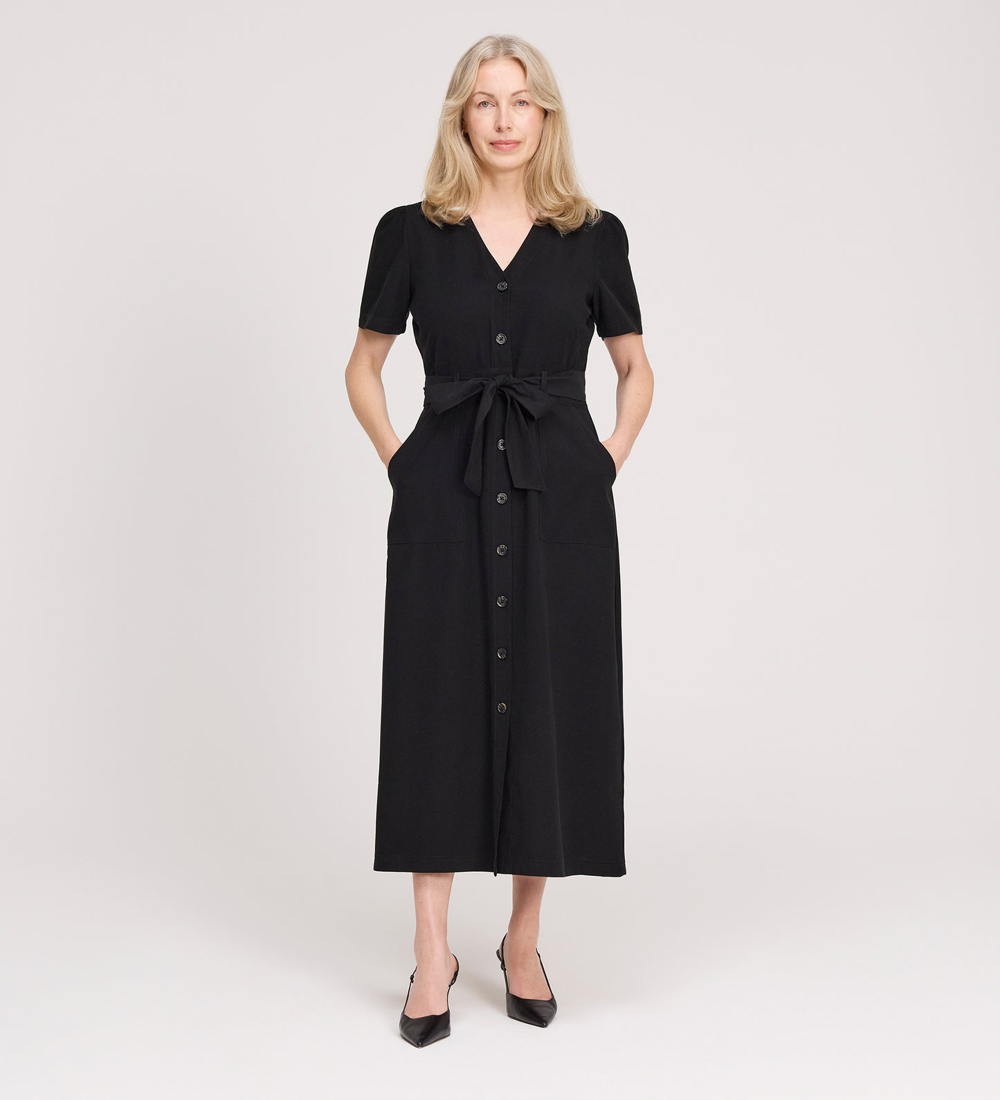 A woman with long blonde hair stands facing forward, wearing the Priscilla Black Cupro Midi Dress with a self-tie belt and black heels, posing against a plain light background.