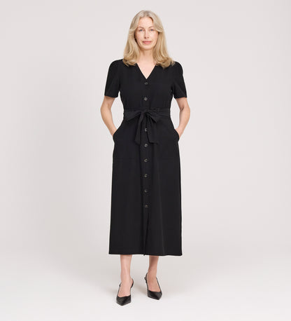 A woman with long blonde hair stands facing forward, wearing the Priscilla Black Cupro Midi Dress with a self-tie belt and black heels, posing against a plain light background.