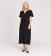 A woman with blonde hair wears the Priscilla Black Cupro Midi Dress with a self-tie belt and short sleeves, paired with black pointed shoes, standing against a plain light background.