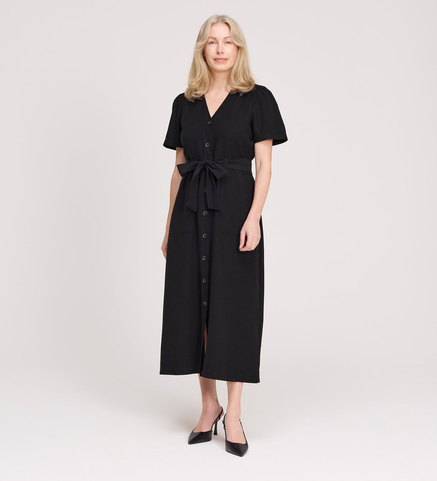 A woman with blonde hair wears the Priscilla Black Cupro Midi Dress with a self-tie belt and short sleeves, paired with black pointed shoes, standing against a plain light background.