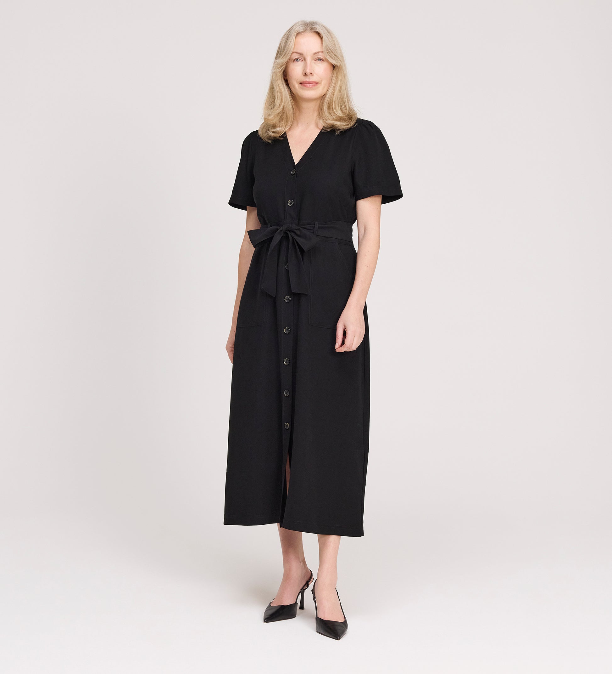 A woman with blonde hair wears the Priscilla Black Cupro Midi Dress with a self-tie belt and short sleeves, paired with black pointed shoes, standing against a plain light background.