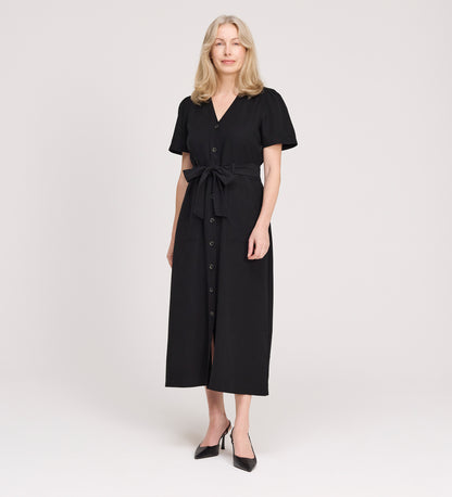 A woman with blonde hair wears the Priscilla Black Cupro Midi Dress with a self-tie belt and short sleeves, paired with black pointed shoes, standing against a plain light background.