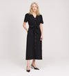 A woman with long, light blonde hair stands against a white background wearing the Priscilla Black Cupro Midi Dress, featuring large patch pockets and a self-tie belt. She smiles slightly, with one hand in her pocket and black heeled shoes.