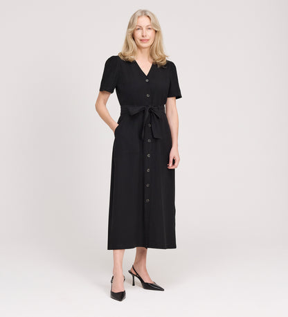 A woman with long, light blonde hair stands against a white background wearing the Priscilla Black Cupro Midi Dress, featuring large patch pockets and a self-tie belt. She smiles slightly, with one hand in her pocket and black heeled shoes.