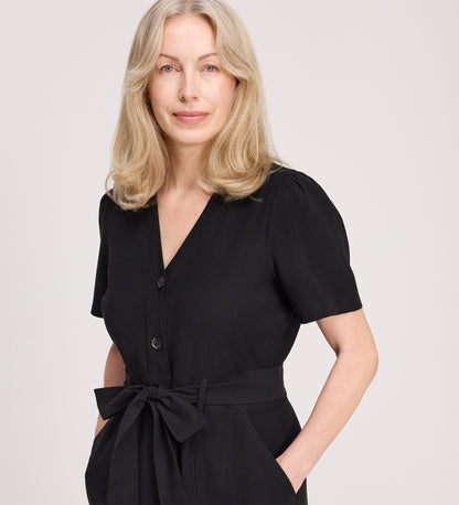 A woman with shoulder-length blonde hair models the Priscilla Black Cupro Midi Dress, featuring large patch pockets and a self-tie belt, standing with her hands in her pockets against a plain light background.