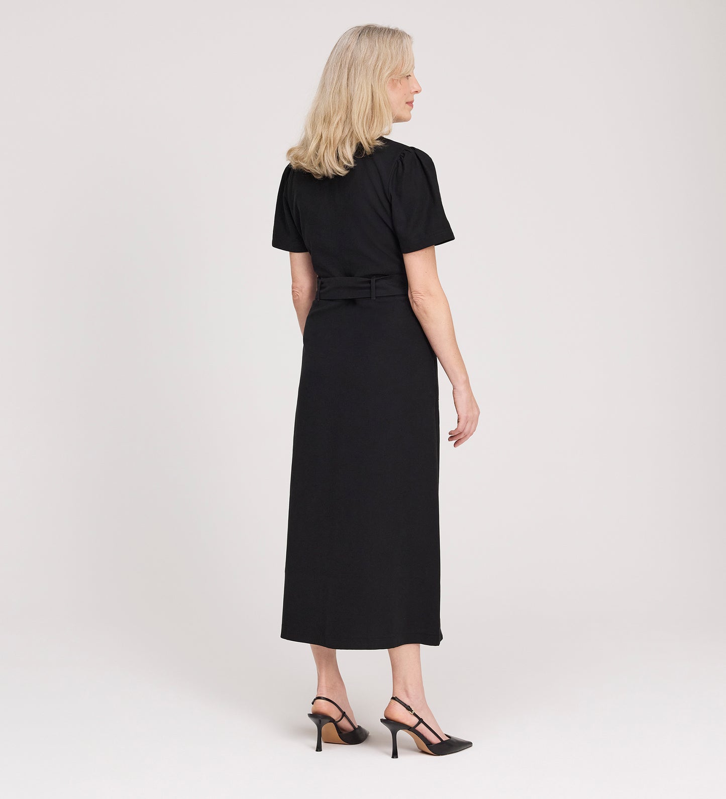 A woman with blonde hair, wearing the Priscilla Black Cupro Midi Dress and black heels, stands facing away from the camera against a plain light background.