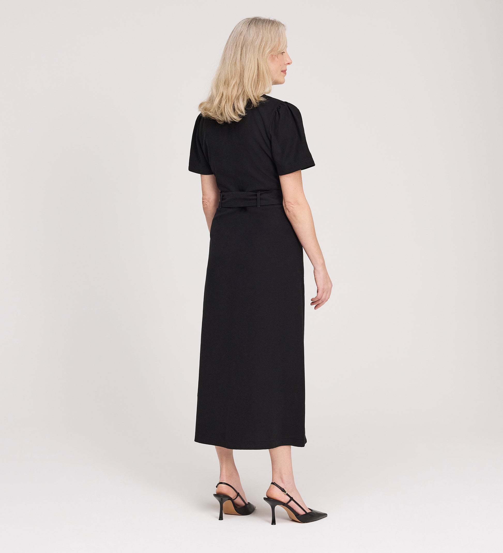A woman with blonde hair, wearing the Priscilla Black Cupro Midi Dress and black heels, stands facing away from the camera against a plain light background.