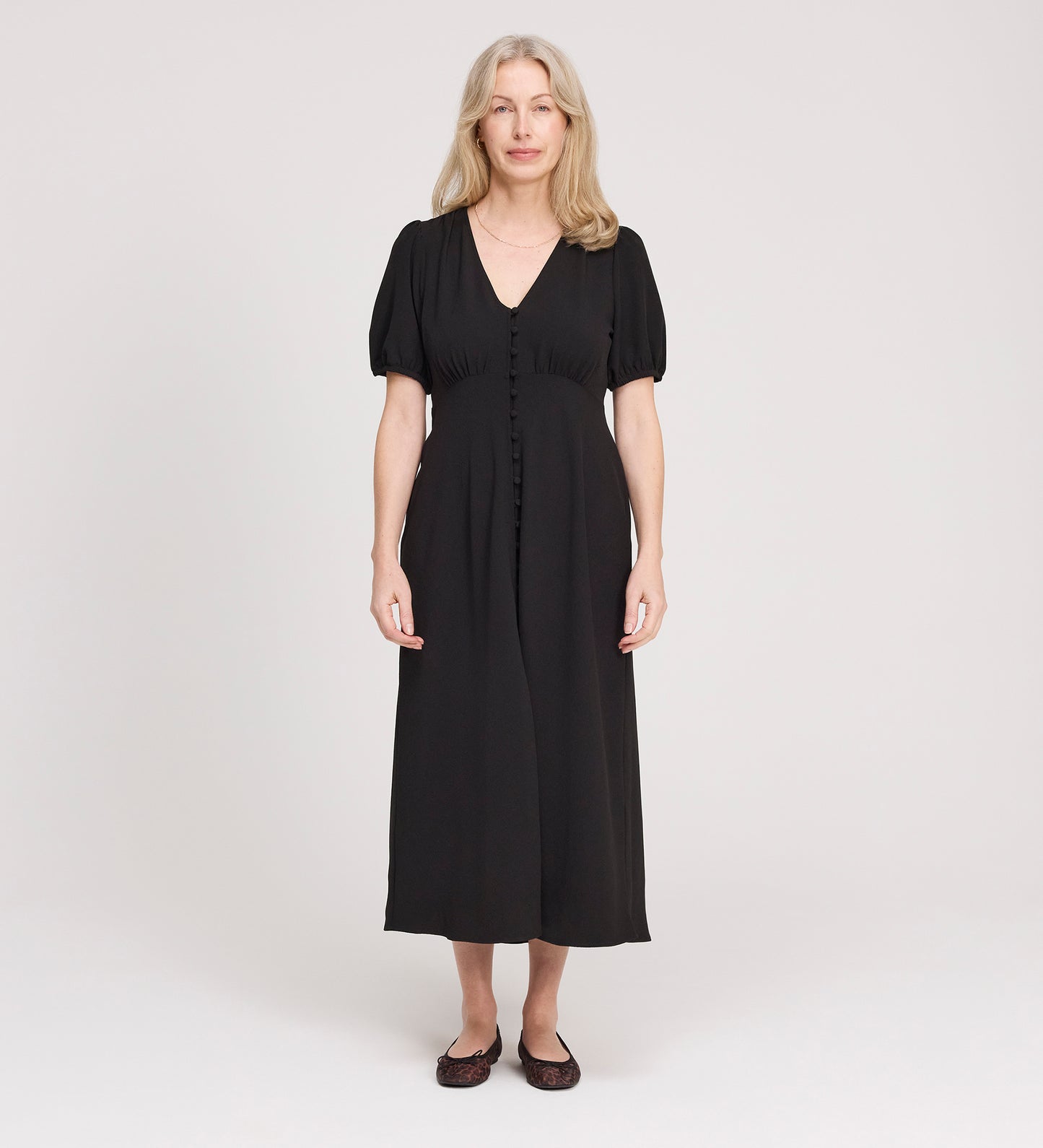 A woman with long, light hair stands facing forward in the Robyn Black Button Through Midi Dress with short sleeves and flat shoes, against a plain, light background. Ideal as a versatile day to evening dress for any occasion.