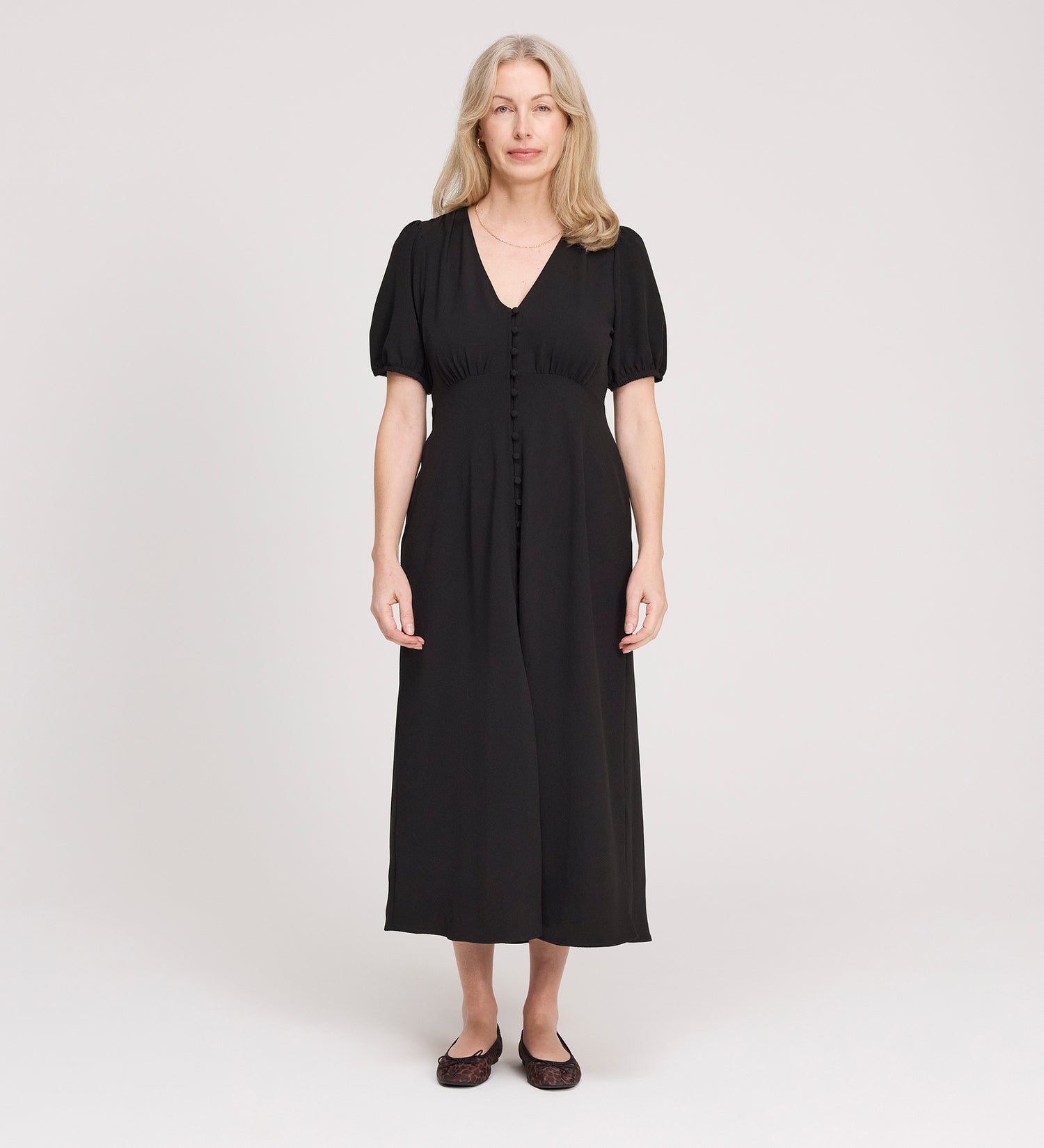 A woman with long, light hair stands facing forward in the Robyn Black Button Through Midi Dress with short sleeves and flat shoes, against a plain, light background. Ideal as a versatile day to evening dress for any occasion.