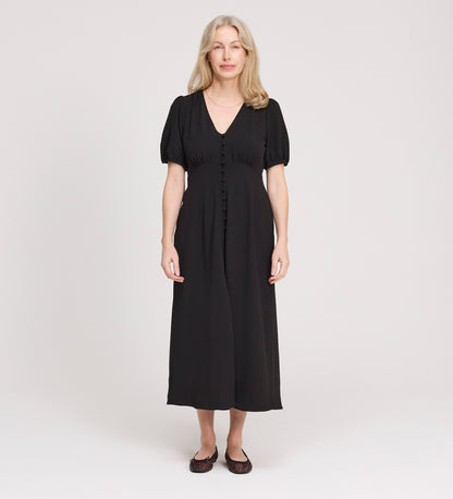 A woman with long, light hair stands facing forward in the Robyn Black Button Through Midi Dress with short sleeves and flat shoes, against a plain, light background. Ideal as a versatile day to evening dress for any occasion.
