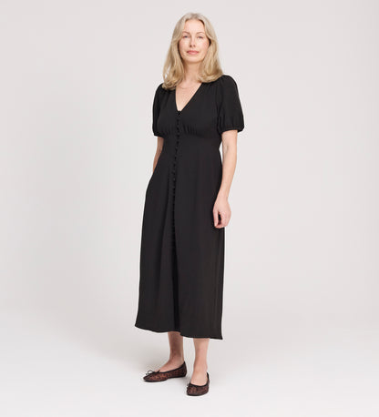 A woman with light hair faces forward and smiles slightly in a studio, wearing the Robyn Black Button Through Midi Dress and dark flats, against a plain light background.
