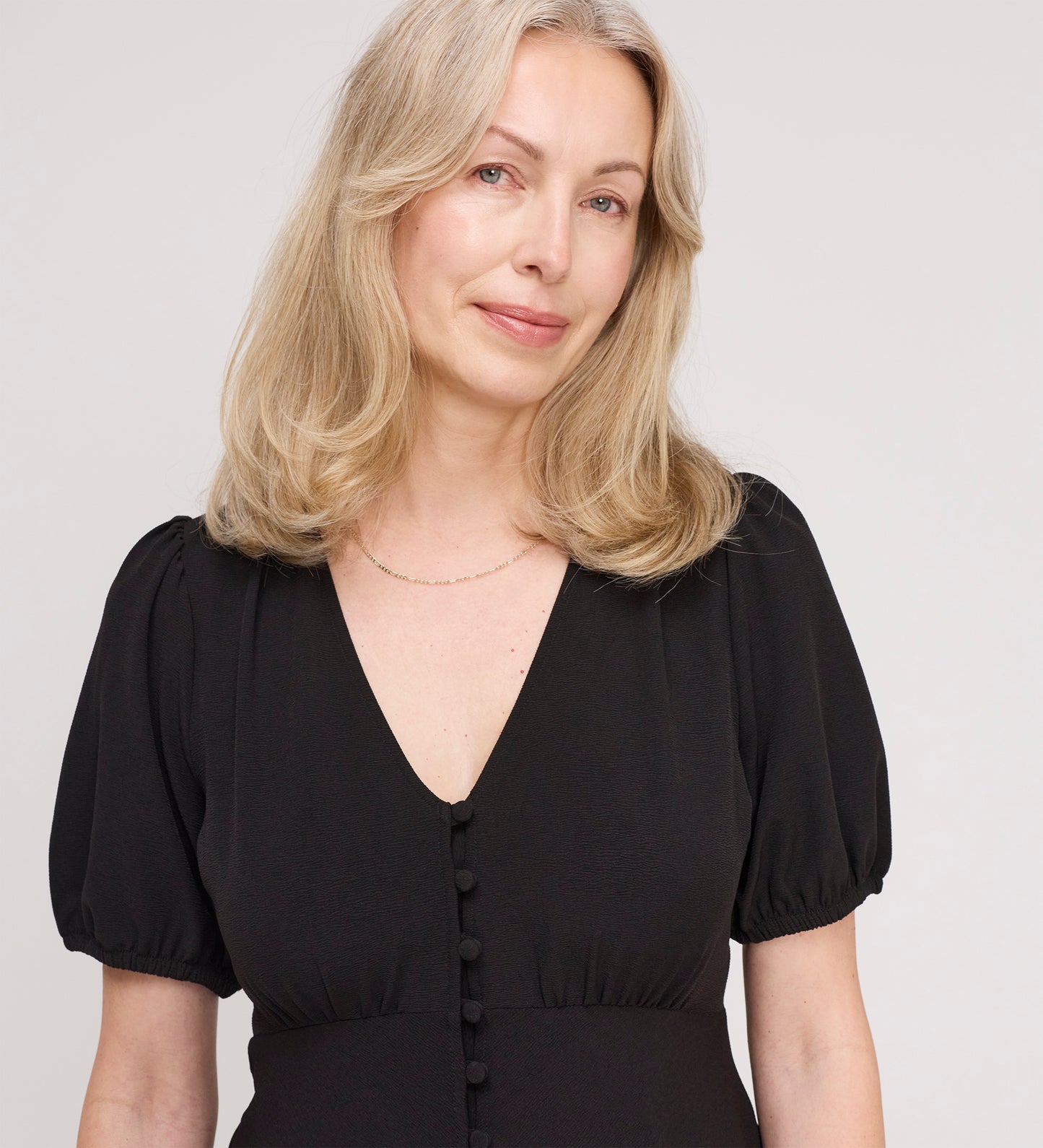 A woman with shoulder-length blonde hair wears the Robyn Black Button Through Midi Dress, featuring an empire waist and V-neckline, standing against a plain light background and smiling softly.
