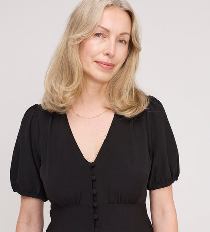 A woman with shoulder-length blonde hair wears the Robyn Black Button Through Midi Dress, featuring an empire waist and V-neckline, standing against a plain light background and smiling softly.