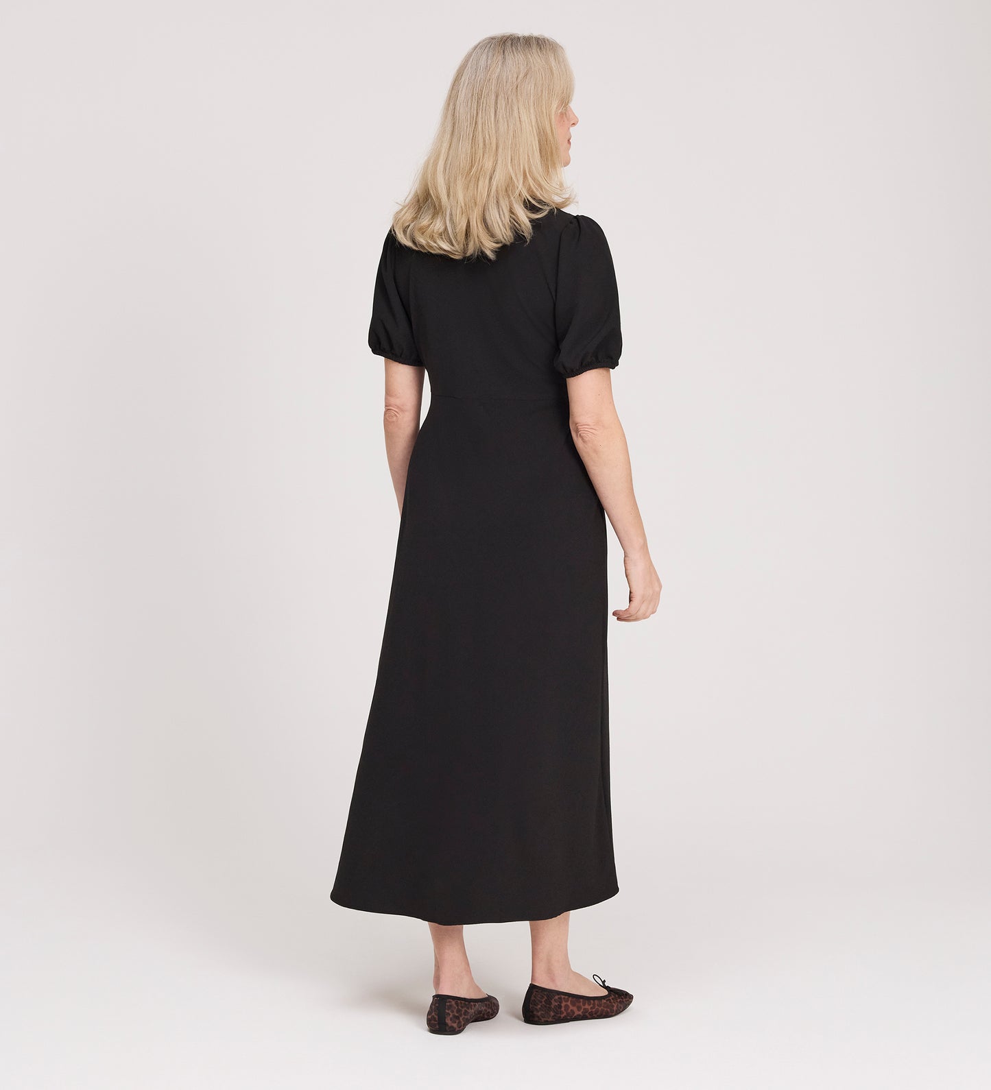 A woman with blonde hair faces away, wearing the Robyn Black Button Through Midi Dress with puff sleeves and black flats, set against a plain light background.