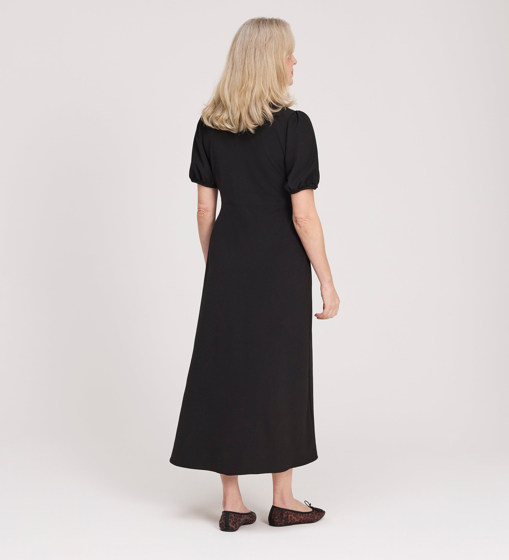 A woman with blonde hair faces away, wearing the Robyn Black Button Through Midi Dress with puff sleeves and black flats, set against a plain light background.