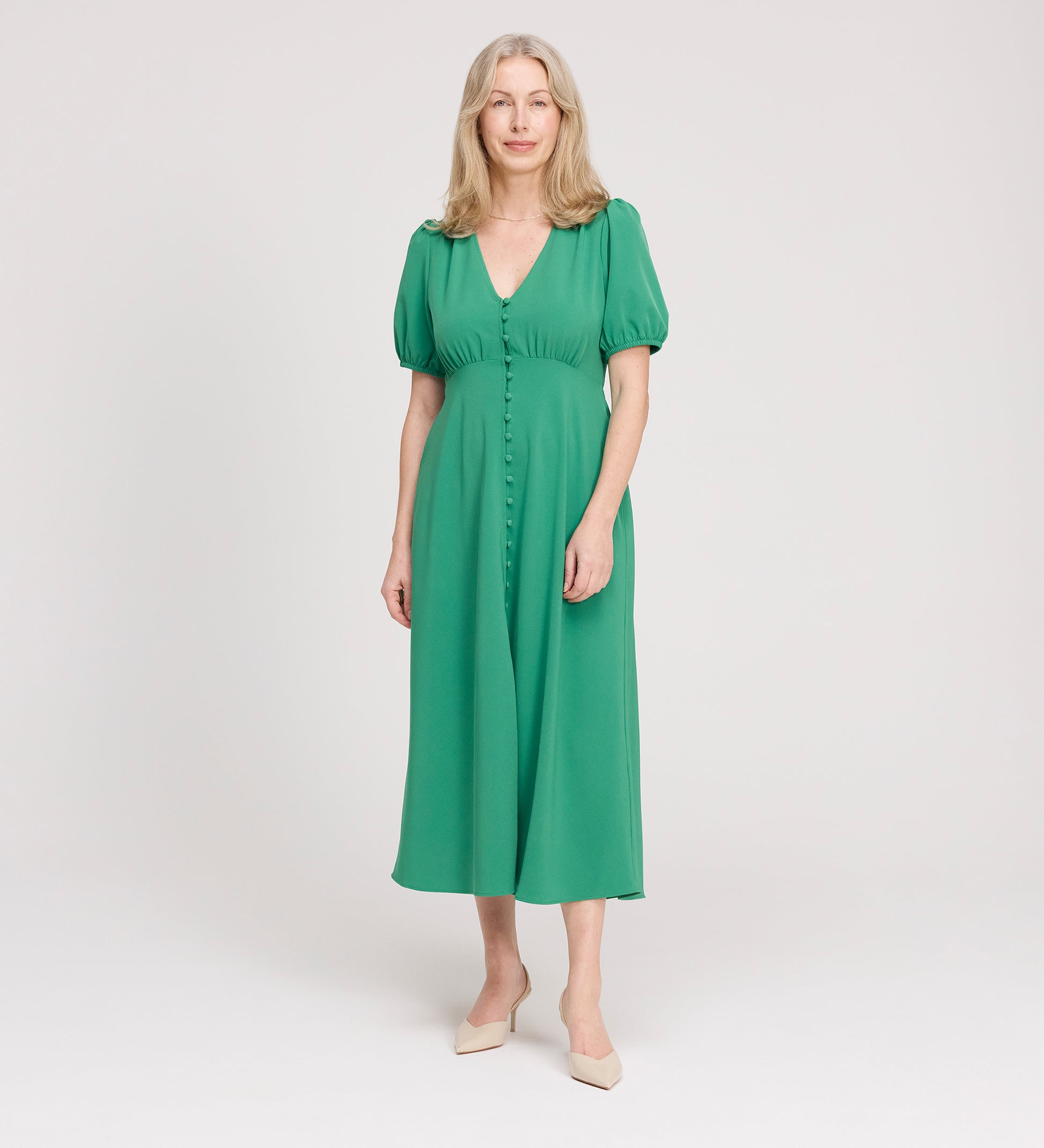 A woman with long, light hair stands against a plain background, wearing the Robyn Green Button Through Midi Dress and beige shoes. She smiles softly with her arms relaxed by her sides.