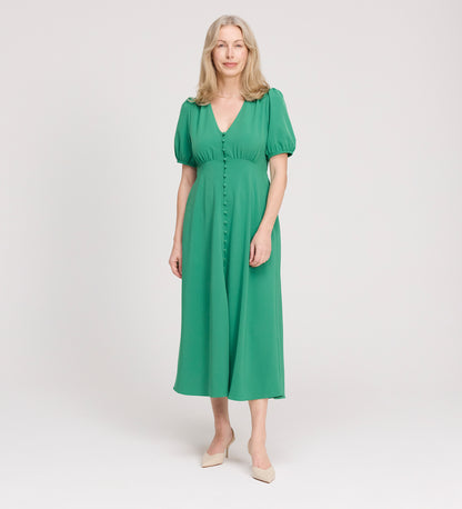 A woman with long, light hair stands against a plain background, wearing the Robyn Green Button Through Midi Dress and beige shoes. She smiles softly with her arms relaxed by her sides.